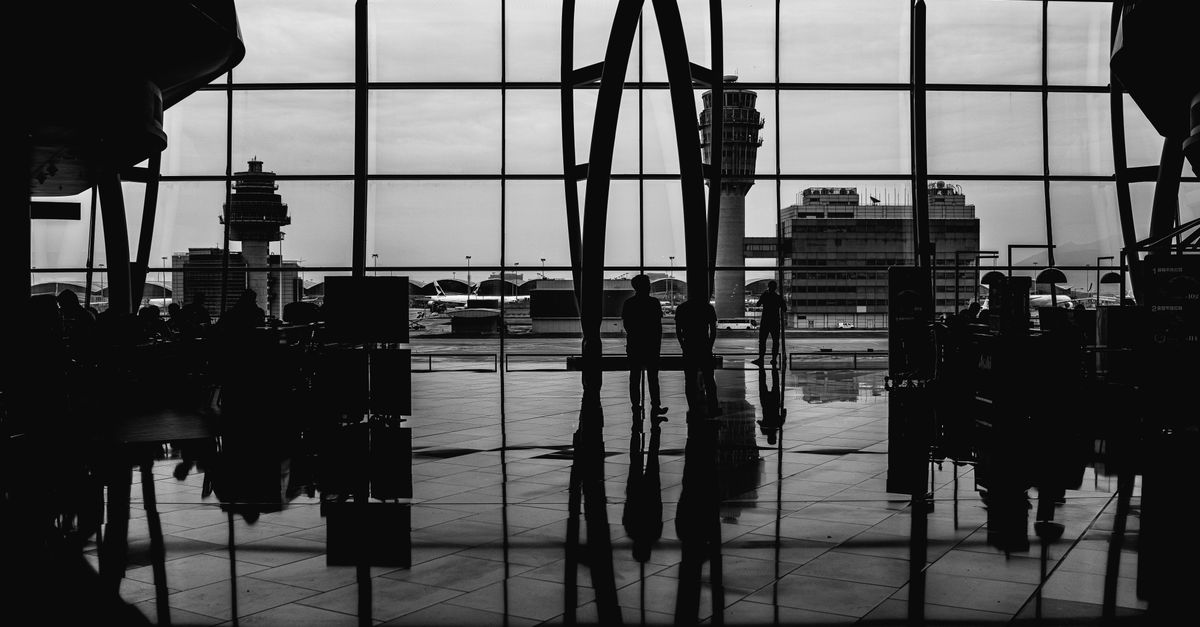 A person standing in an airport terminal, looking out of a large window at a control tower and another building.