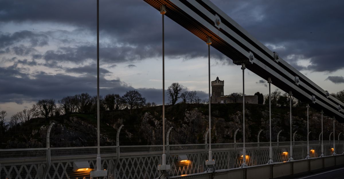 A scenic twilight view of a historic suspension bridge in the UK with a castle in the background.