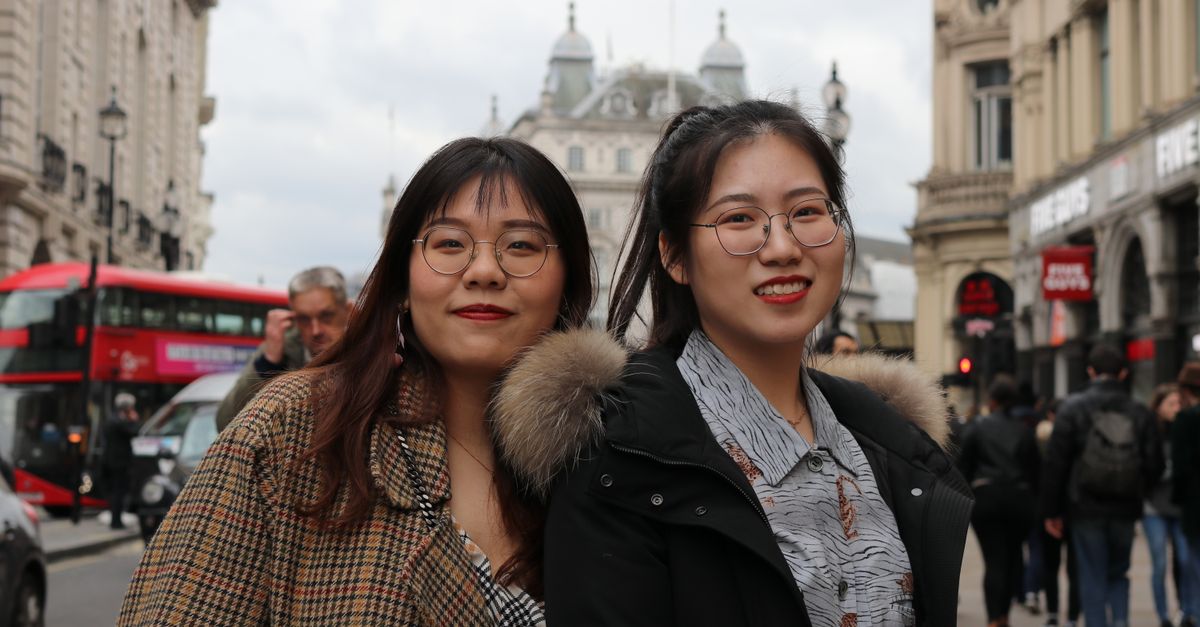 Two friends smiling on a busy street in London, with iconic architecture and a red double-decker bus in the background.