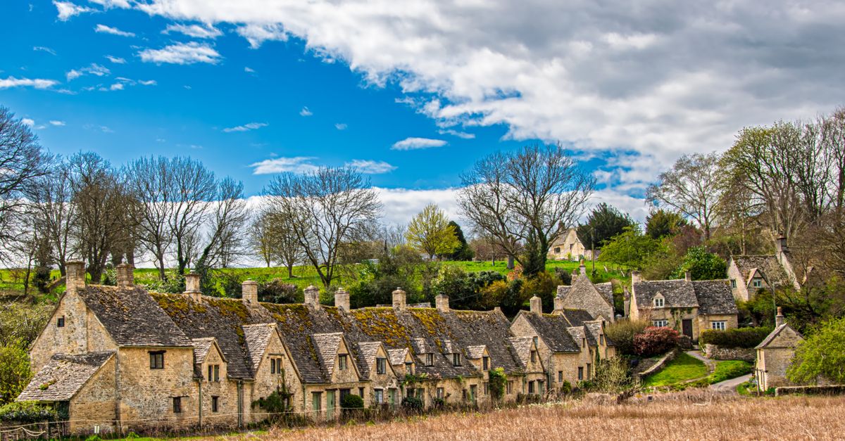 Row of honey-stone Cotswold cottages with mossy roofs beneath a blue sky