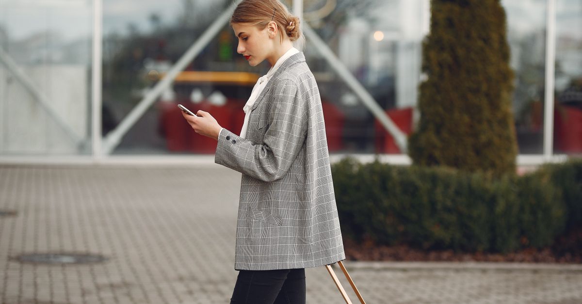 A traveller checking her phone while walking through a terminal with a wheeled suitcase.