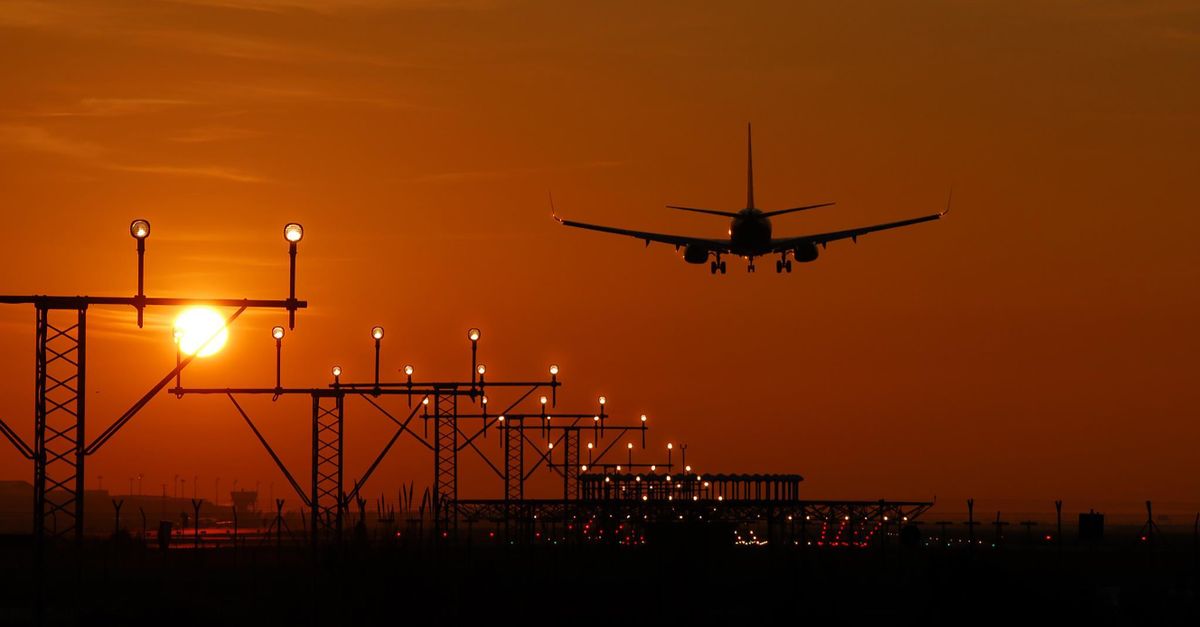 A plane landing at sunrise, silhouetted against a glowing sky, approach lights below.